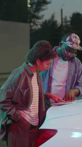 Vertical side full medium shot of African American fellow road trippers deciding on route studying country map on car hood at night Royalty-Free Stock Footage #3457212125