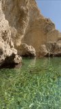 Vertical video, Panorama of beautiful rocky bay in Mediterranean Sea at Stegna beach, Rhodes island, Greece. A woman standing in water inspects coastal rocks, slow motion