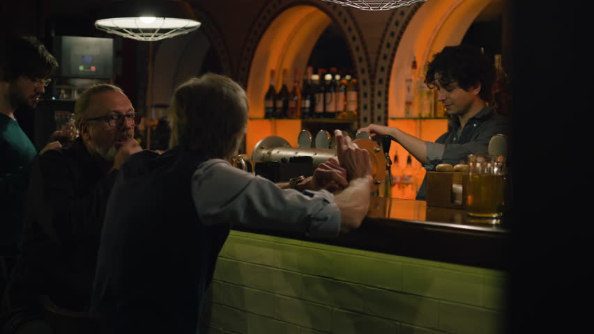 Young bartender pours craft beer into glass and gives to mature man. Two male friends drink beer, talk to barman sitting at the bar counter in stylish pub at night. Concept of lifestyle and leisure.