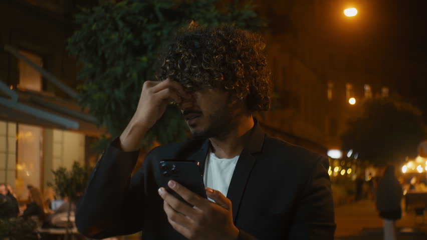Indian Arabian Latino man holding mobile phone indignant confused angry displeased mad ethnic male cellphone user technology city outside night negative trouble problem spam furious notification lost Royalty-Free Stock Footage #3457243763