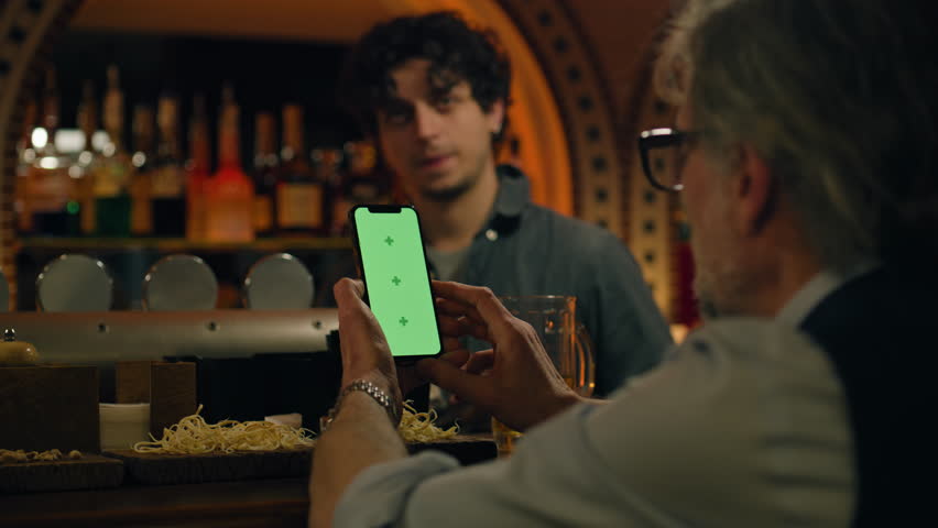 Male sports fan uses mobile phone with green screen, bets on football match online, talks with bartender and friend. Mature man checks bookmaker ratings in app sitting at bar counter in pub. Mock-up.