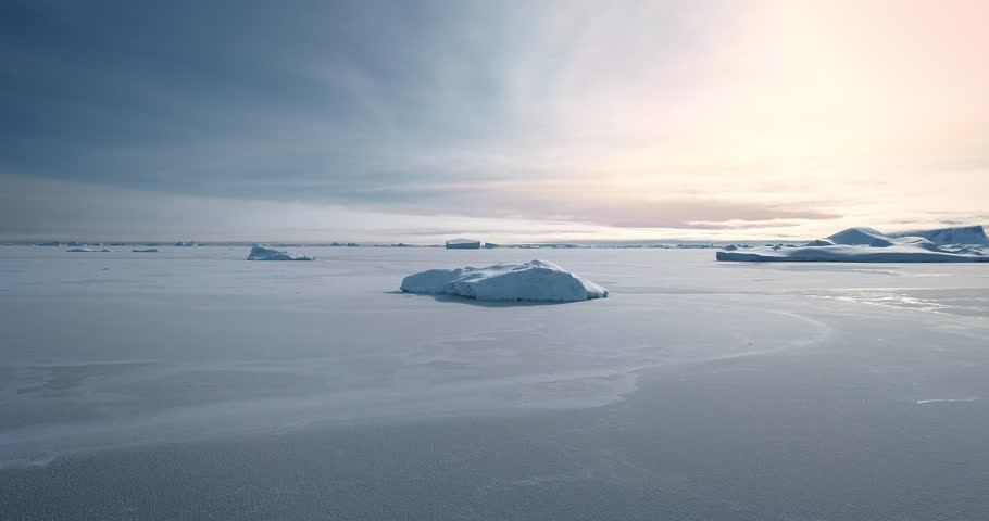 Breathtaking journey over frozen Antarctica landscape. Desert white land of snow and ice aerial drone shot. Several blue icebergs on ocean surface. Sunset sky. Discover the beauty of South Pole