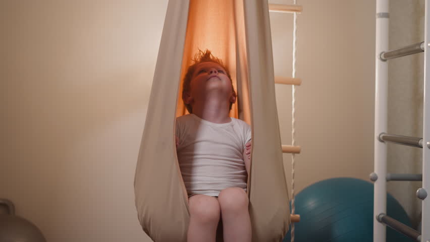 Curious blond boy swaying in comfortable beige hammock looking at ceiling. Cute little child in t-shirt enjoys relaxing in modern home gym slow motion