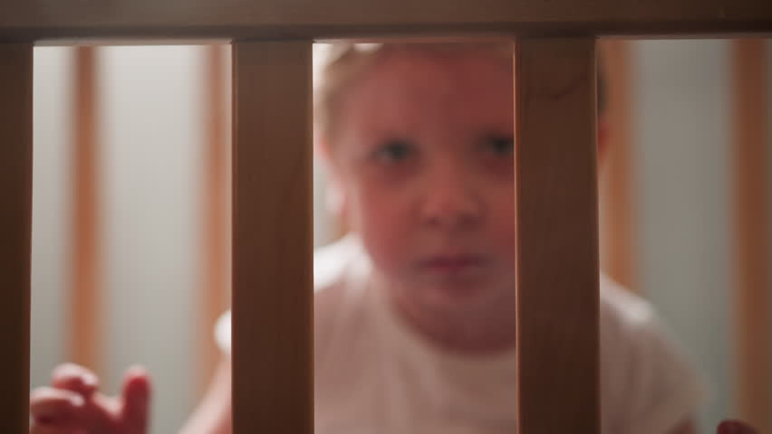 Upset blond toddler looks out of cot grabbing wooden planks with hands. Scared little boy looks ahead sitting in nursery alone closeup slow motion Royalty-Free Stock Footage #3457370753