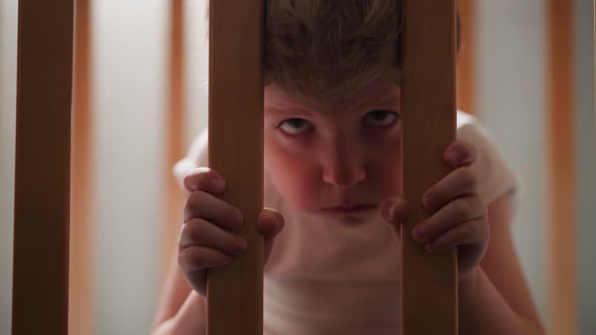 Offended little boy looks through wooden grid of baby cradle in children room closeup. Upset toddler kid hides in bed. Child suffers from loneliness Royalty-Free Stock Footage #3457372727