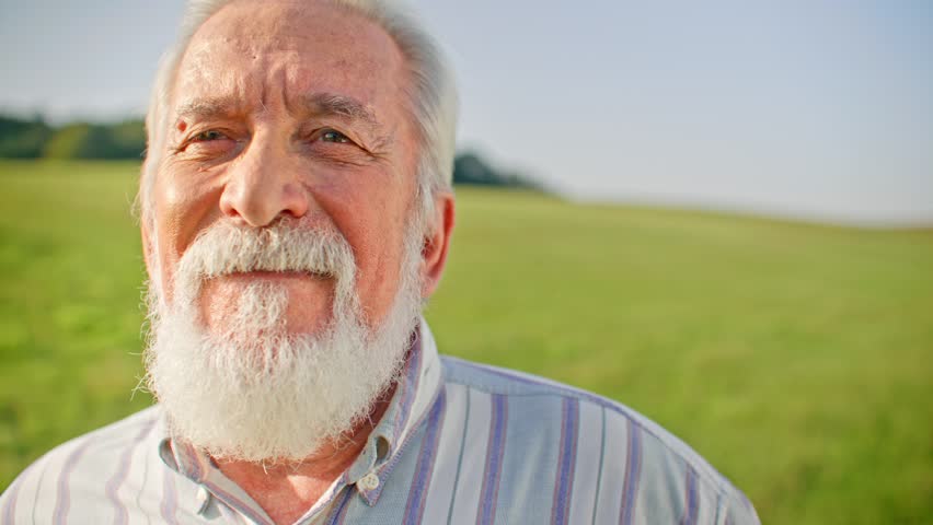 Portrait of older Caucasian man looking past camera and smiling. Large green field in background with forest on horizon. Male with white beard. Happy man. Concept of positivity.