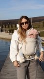a mother in sunglasses stands with a child in a sling against the background of a lake and walks with a dog. vertical video