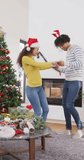 Vertical video of happy diverse couple in christmas antlers and santa hat dancing at home. Christmas, celebration, happiness, tradition and inclusivity concept.