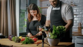 Young happy couple in an apron standing in kitchen records on smartphone food videoblog. Couple cutting vegetables, preparing healthy a salad. Blogging, healthy lifestyle, cooking master class