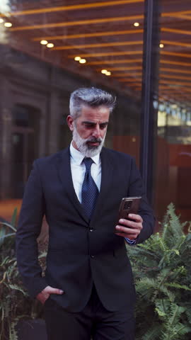 Mature businessman in suit reads message on his smartphone, standing outdoors near modern office building, embodying professionalism and focused attention. Successful executive using banking app