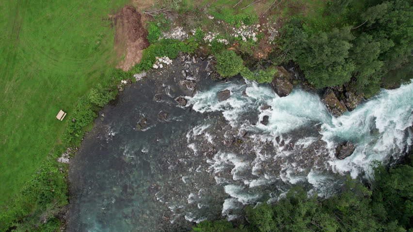 Top down aerial view, tracking over a fast flowing river in Norway. White water rapids form as it crashes over large rocks. The waterfall flows into a wide, calm pool. Dense forest grows on both sides