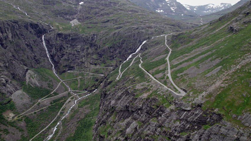 Wide aerial shot of the Trollstigen in Norway. Multiple tight hairpins weave their way down the steep rocky valley sides, crossing repeatedly in front of the tall Stigfossen Waterfall. Royalty-Free Stock Footage #3457530427