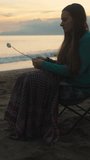 A young woman at sunset by the sea, frying marshmallows on a bonfire. It's cold, she's covered with a blanket. Vertical video Social Media