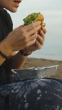 A young woman on the seashore, with her mouth wide open, bites off a large piece of burger. Picnic by the sea. Vertical video Social Media