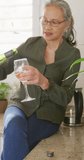 Vertical video of happy asian senior woman drinking wine in kitchen. Active retirement and lifestyle concept.