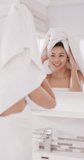 Vertical video of smiling biracial woman with towel on hair looking in mirror in bathroom. Health and beauty, leisure time, domestic life and lifestyle concept.