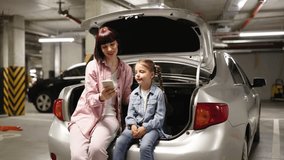 Caucasian family having video call using cell phone sitting on the trunk of car before exciting trip. Caucasian mother and preschool daughter making selfie with smartphone in underground parking lot.