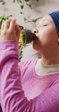 Vertical video of smiling biracial woman in hijab drinking water from bottle after workout at home. Happiness, health, fitness, inclusivity and domestic life.
