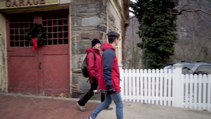 Teenage boy vlogger with camera and mature mother walk down a sidewalk in winter in Harpers Ferry, West Virginia.
