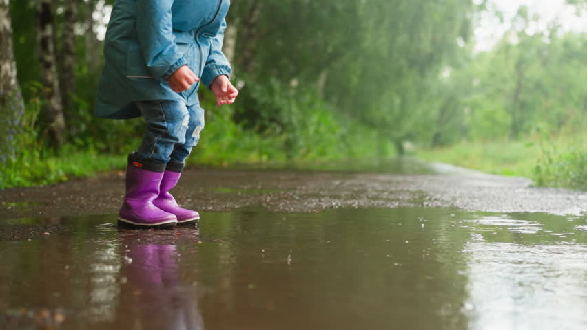 Little child jumps in puddle on park road closeup. Funny little boy plays splashing water water on rainy spring day. Outdoors activity and happy childhood