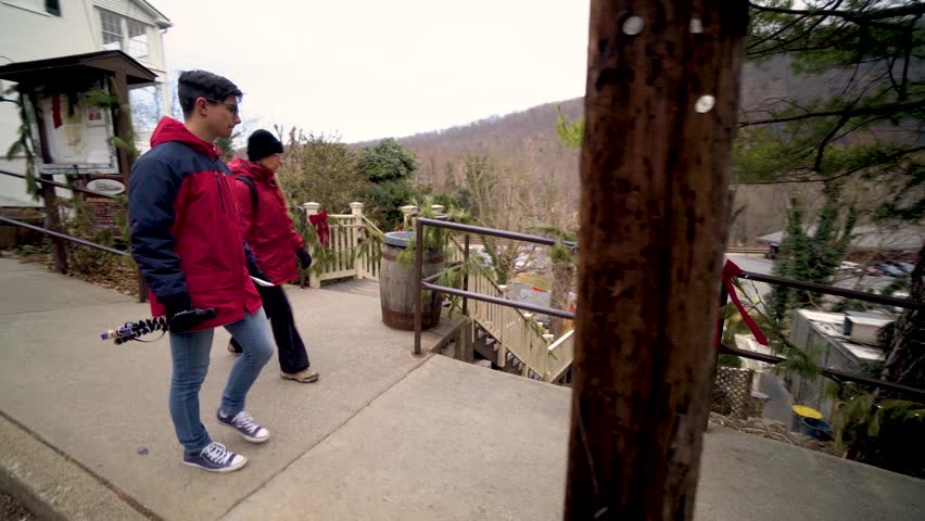 Teenage boy vlogger with camera and mother walk down a brick sidewalk with Christmas decorations on railing in Harpers Ferry National Park, West Virginia.