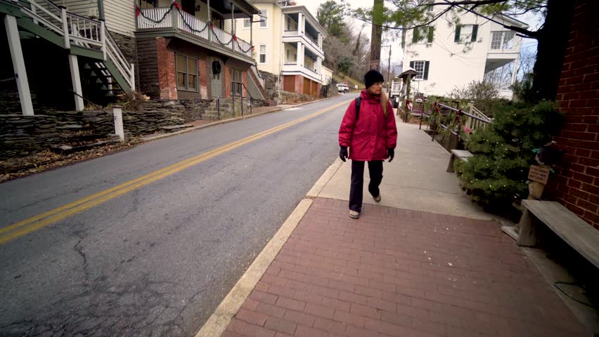 Mature woman walks down a sidewalk and waits for her teenage vlogging son to photograph in Harpers Ferry, West Virginia.