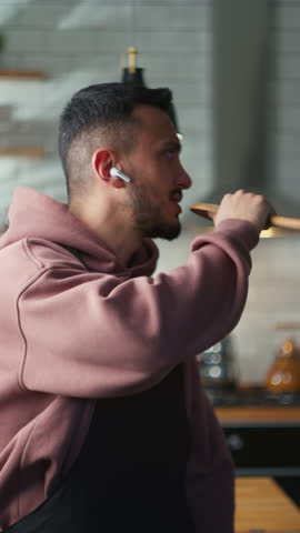 Vertical video. Carefree man with wireless earphone singing into a ladle like a microphone and freely dancing to music in a kitchen at home