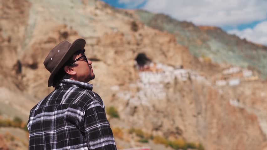 Rack focus shot of an Indian male tourist wearing hat looking at the Phugtal Monastery in remote area of Zanskar Valley in Ladakh, India. Focus changes from Man to the Monastery in background.