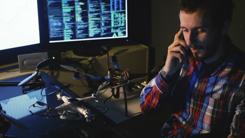 Young bearded Male Engineer work with drone in workshop. Computers and monitors background. 