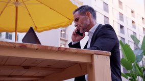  Businessman talks on a smartphone while working with laptop outdoors.