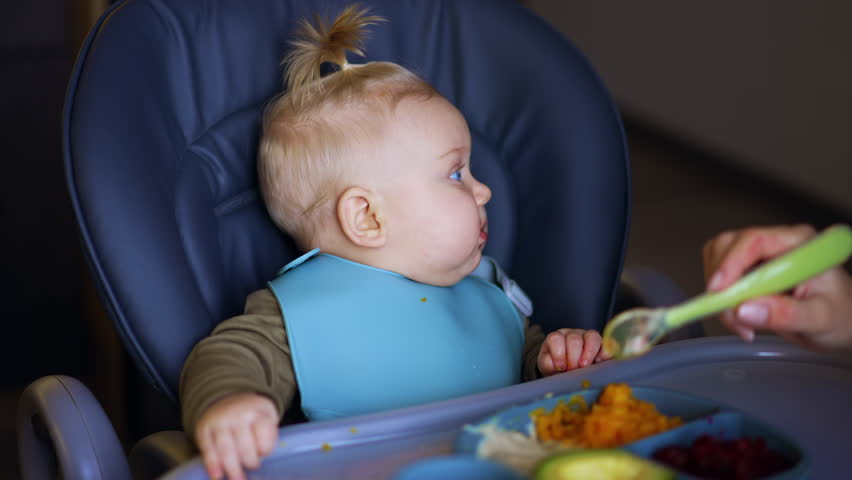 Blue-eyed blond baby is given food from spoon. Mother feeding her child with healthy vegetables.