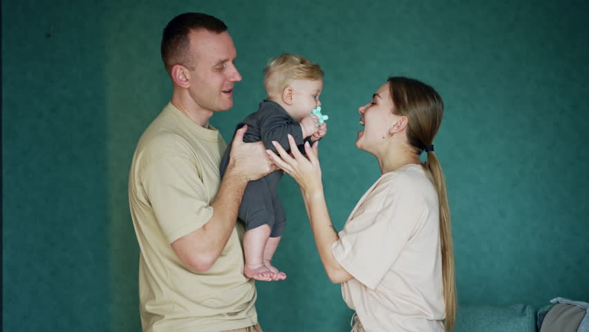 Young Caucasian parents are playing with their little baby. Man holds a kid and mom teasing a child with a toy.