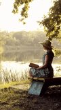 Beautiful young woman in dress and hat sitting on bench under tree and reading book at sunset. Lady with book resting relaxing on nature near lake or river outdoors. Enjoying vacation vertical video