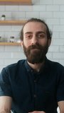 Vertical video. Young bearded man sitting in modern kitchen at home talking to camera making conference business call. Video call event by work or study concept
