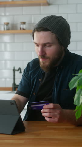 Vertical video. Anxious man with beanie sitting in kitchen at home takes his credit cards out of his pocket and tries them one by one to find the credit card that has not reached its limit, gives up