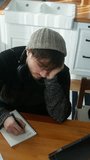 Vertical video. High angle shot of young student man with beanie sitting in modern kitchen tears the page from notebook and throws it back while study with tablet computer and takes notes in notebook