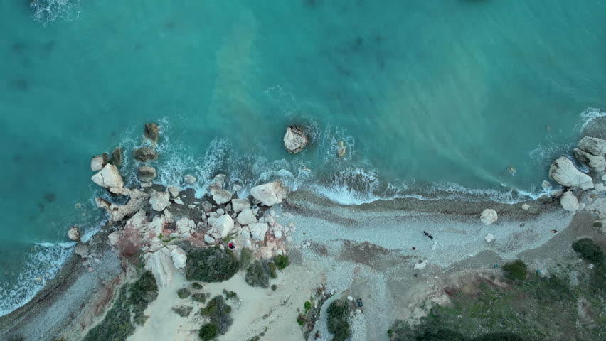 Aerial perspective of a rocky beach with a mixture of sand and boulders, clear blue sea water washing ashore, with visible underwater rocks near the coastline. Royalty-Free Stock Footage #3458120089