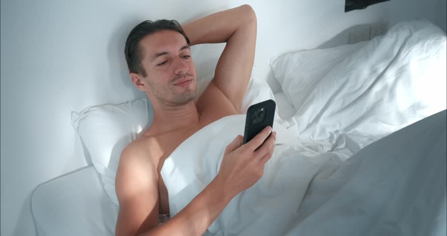 Awakened man lying in bed using phone window to world's news Awakened man scrolls phone syncing with day's pulse. Awakened man strategizes his morning ritual setting stage for success. Royalty-Free Stock Footage #3458184617