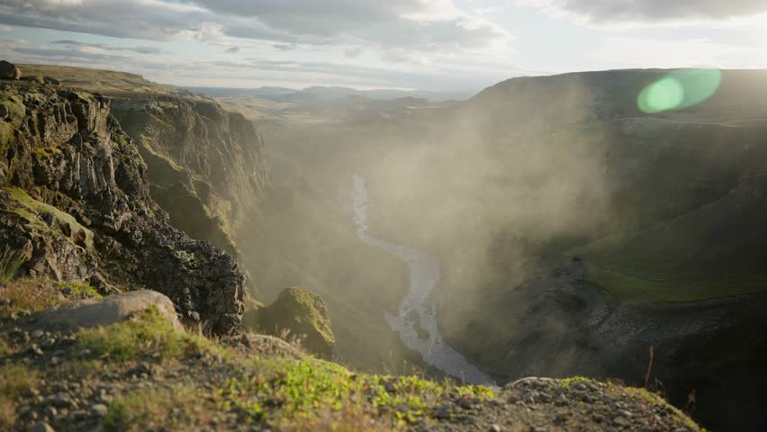 Breathtaking cinematic panoramic shot of green cliffs with a river running through the center. Majestic and beautiful scenery is highlighted by the mist of water in the air. Iceland. Scandinavia.