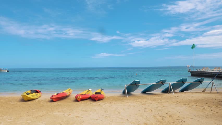 4k Slow motion Video A stunning beachfront scene in Ocho Rios, Jamaica, features surf boards and catyaks dotting the shore, with crystal-clear blue waters and a vibrant blue sky as the backdrop.