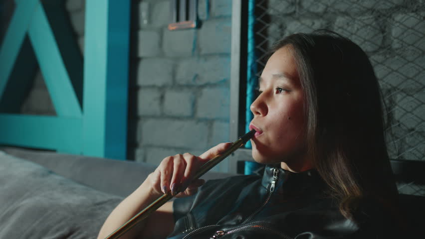 Asian young woman takes of mouthpiece to her lips, inhales and blows out puffs of white smoke at shisha cafe. Pretty adult girl smoking hookah sitting at couch at bar. Female smoker. Side view