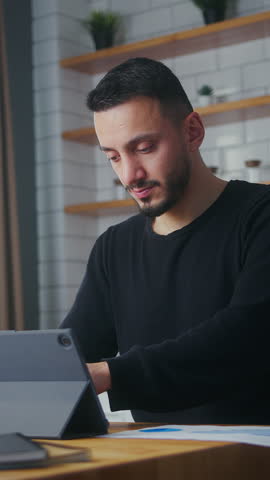 Vertical video. Businessman working at home in the kitchen with his tablet computer, taking notes, doing paperworks. Adult freelancer working on modern tech device