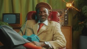Handsome African-American man in stylish outfit sitting on chair, holding vintage telephone and looking at camera with confident smile in retro studio. Video portrait