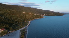 Drone flight along the coast in early morning Greece, showcasing villages and mountains. Morning sun illuminates the mountains and villages. Road runs along the coast.