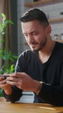 Vertical video. Adult man playing mobile game on the smartphone while sitting in kitchen at home. Excited man engrossed in interesting game, doing winner gesture