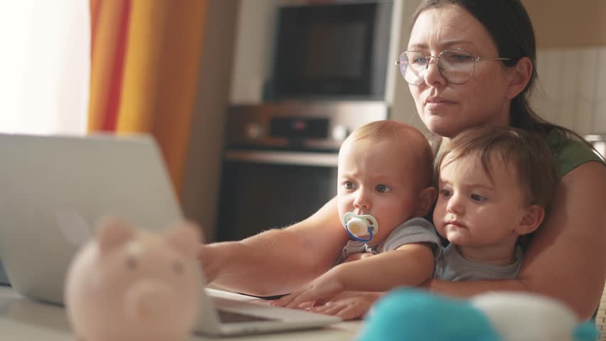 mother works remotely from home with two babies in her arms. pandemic remote work concept. mom tries to work at home in fun kitchen, small children interfere with sitting in her arms business Royalty-Free Stock Footage #3458354409