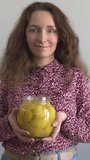 A young, smiling woman in a dress with a white and burgundy pattern presents a jar of homemade preserved lemons. Vertical video.