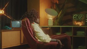 Handsome African-American man with curly hair wig sitting in chair, turning to camera and posing with confidence in studio with vintage interior. Video portrait, retro glow effect