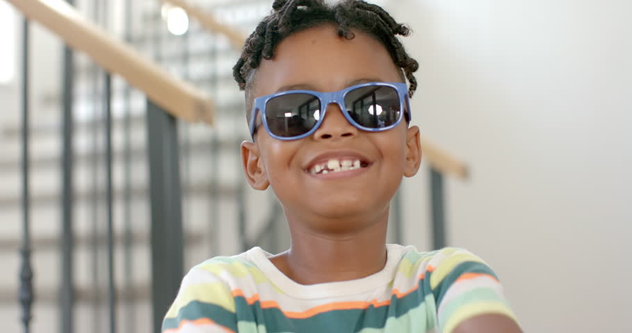 African American boy with a bright smile wears blue sunglasses and a striped shirt at home. His curly hair is styled in small twists, and he exudes joy in a home setting, slow motion.