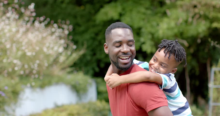 African American father gives his son a piggyback ride in a sunny park at home with copy space. Both are smiling, enjoying a playful and affectionate moment outdoors, slow motion.